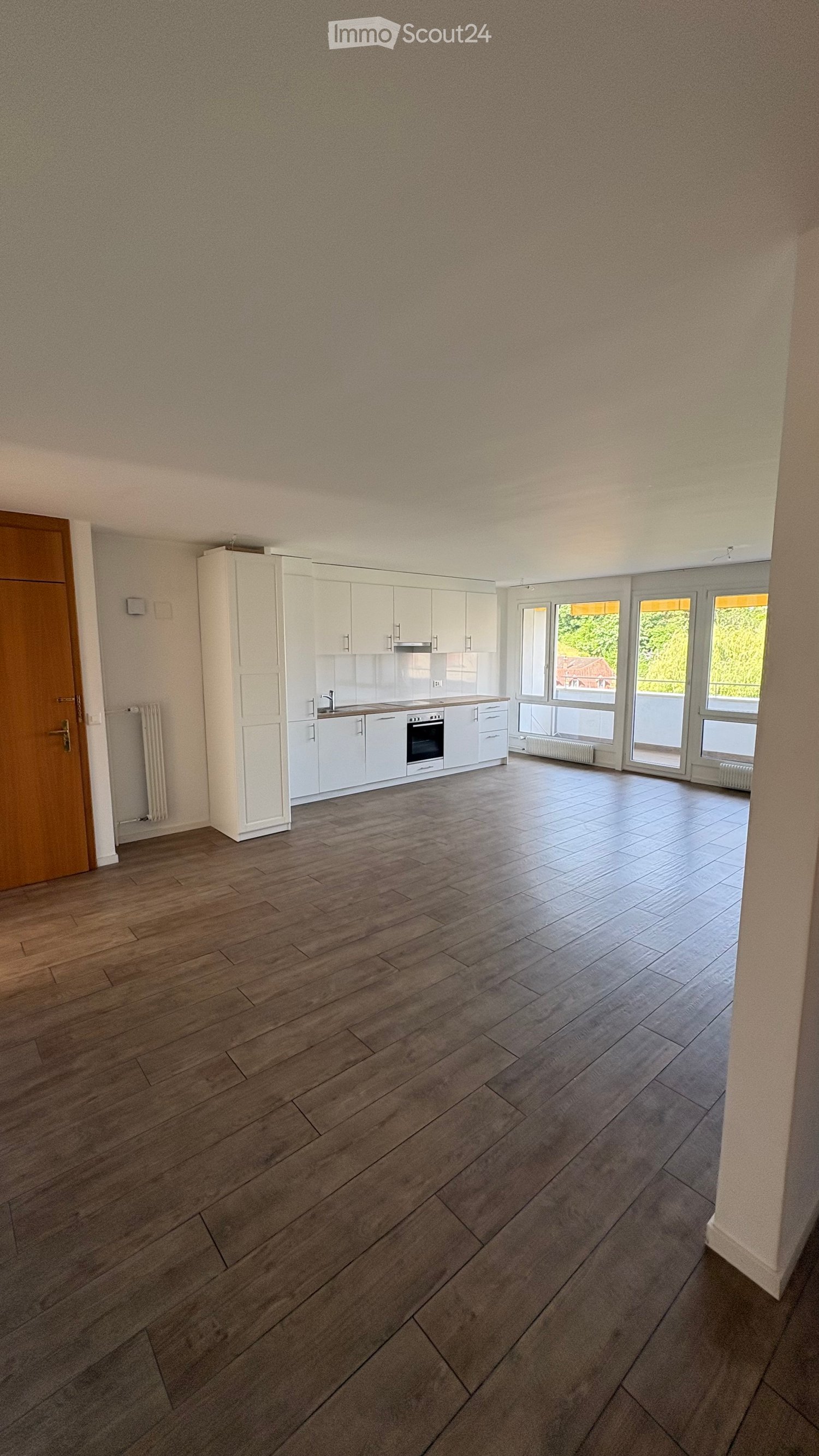 Empty kitchen with white cabinets, wooden floor, and large windows overlooking a green area.
