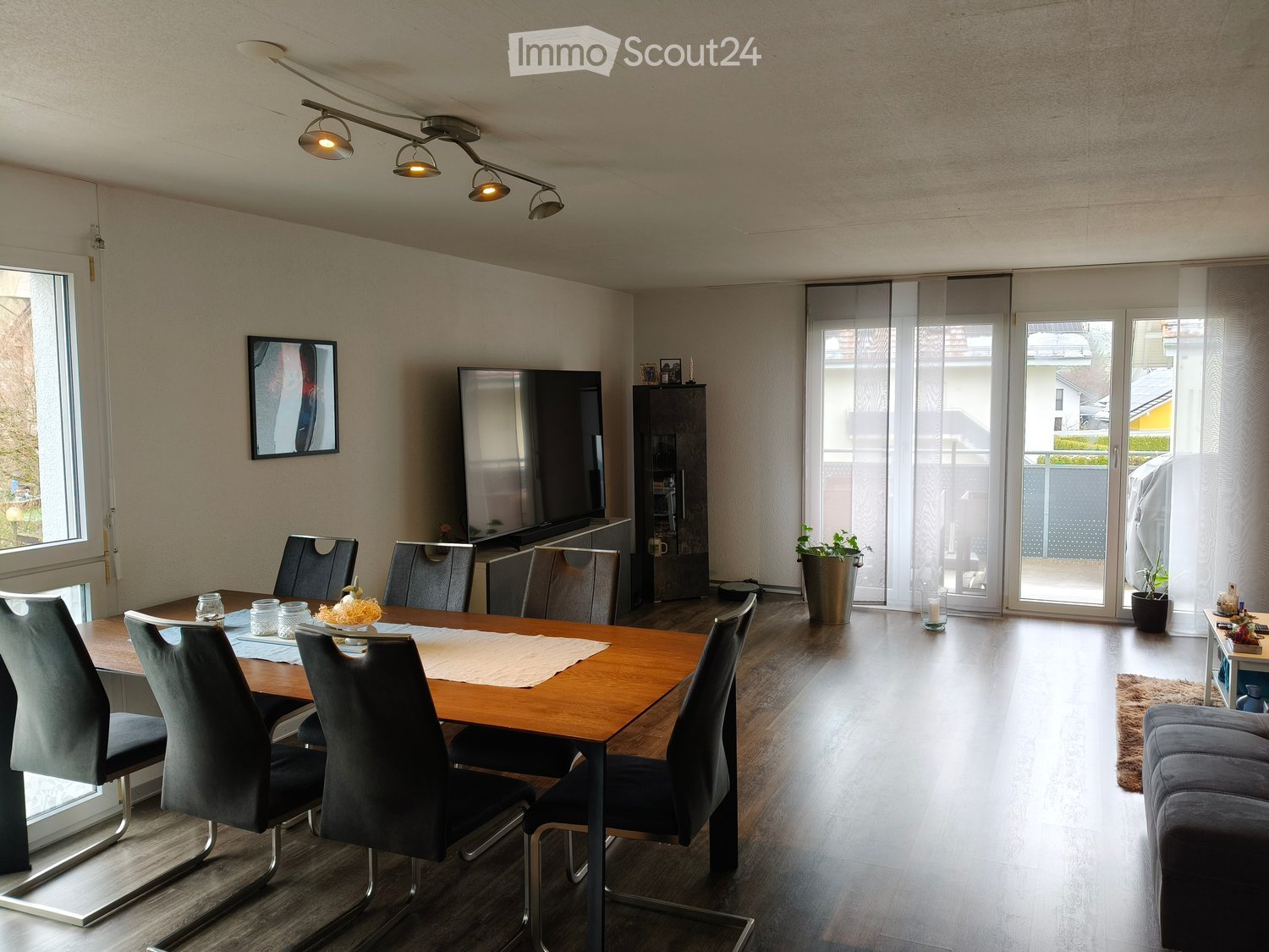 Modern dining area with a wooden dining table, six chairs, a television, a cabinet, and a potted plant near the sliding glass doors.