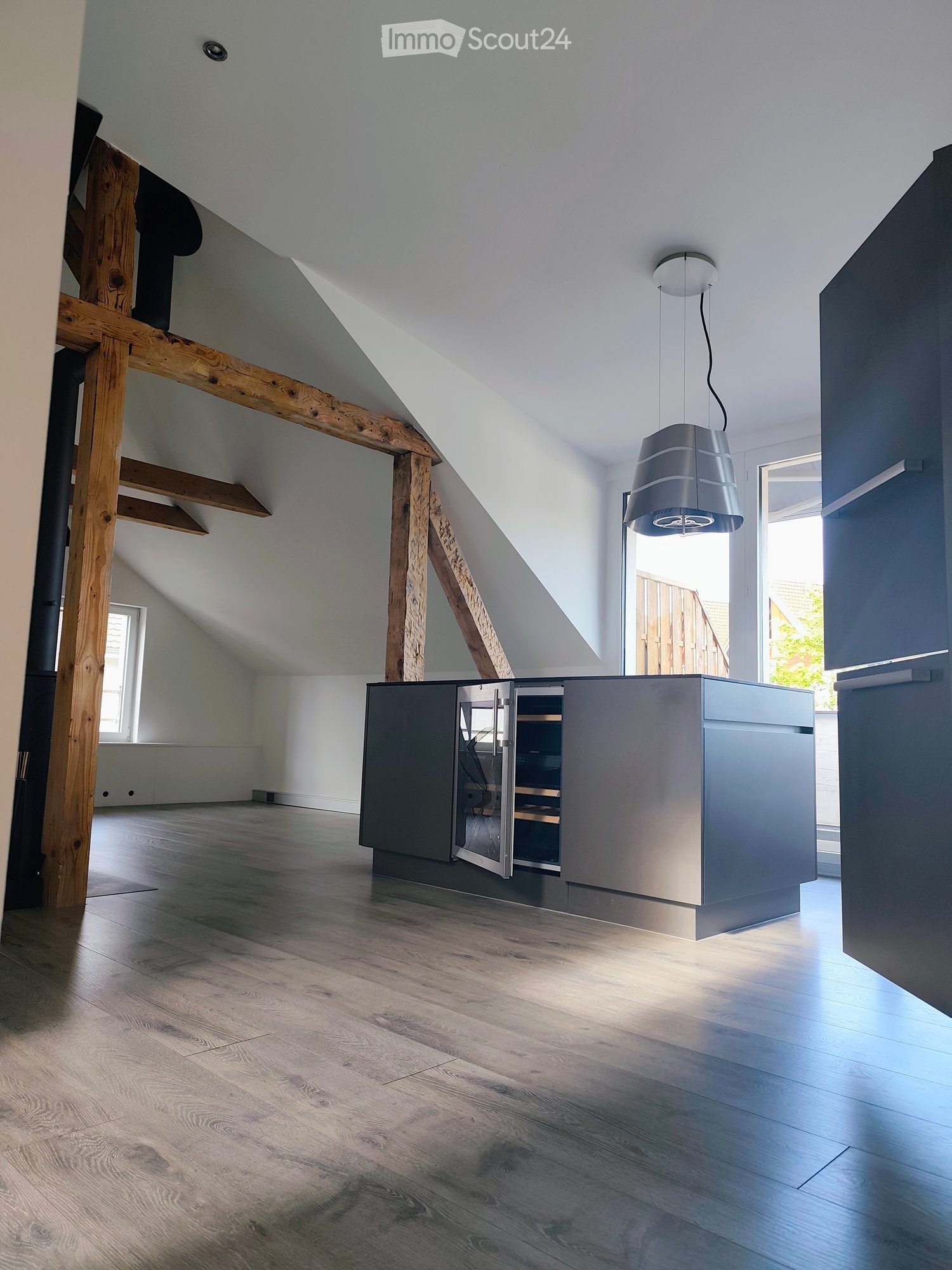 Modern kitchen with sleek grey cabinets, wooden flooring, and a hanging pendant light. The space features exposed wooden beams and large windows allowing natural light to fill the room.