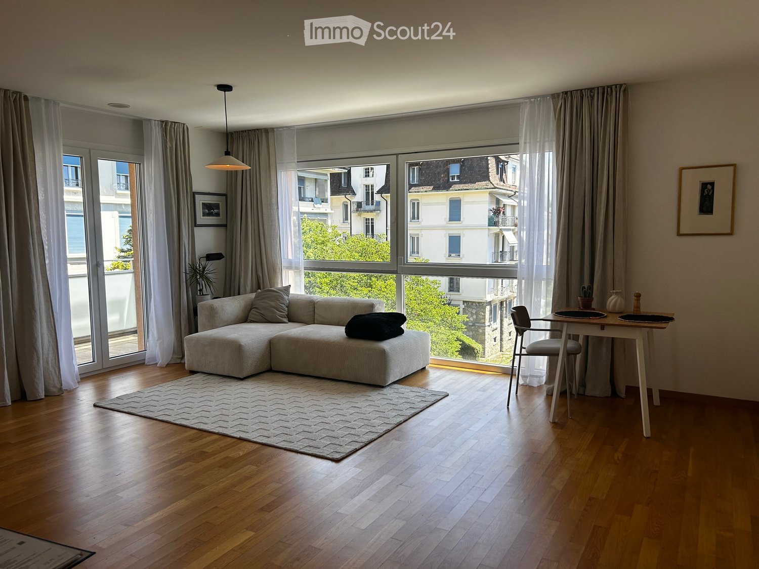 Modern living room with large windows, wooden flooring, a sectional sofa, a coffee table, a dining table with chairs, and decorative items.