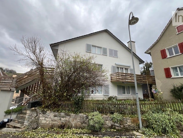 Two-story house with white exterior, multiple windows, raised ground floor, and a wooden balcony on the upper level.
