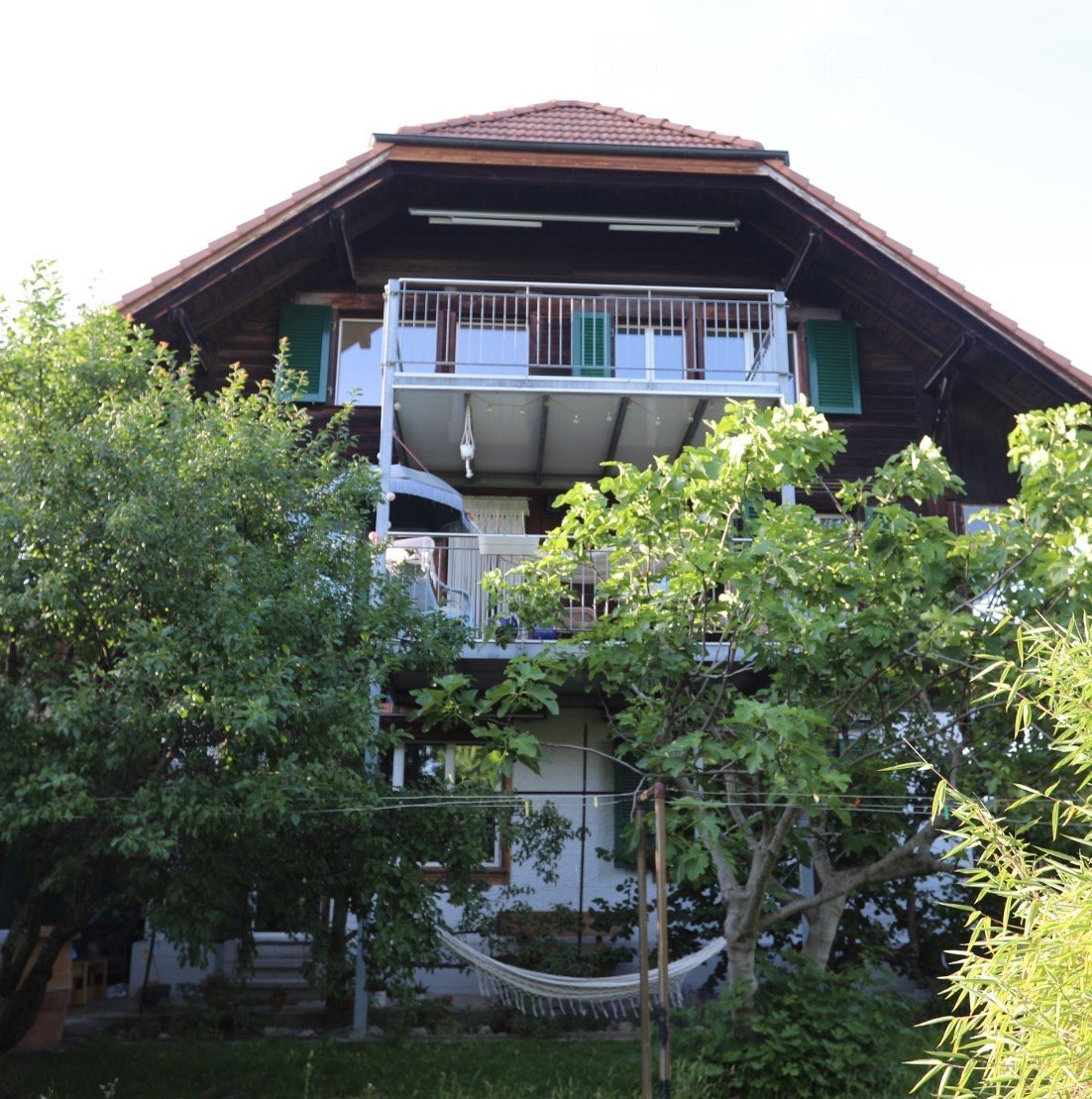 Two-story wooden house with a balcony, green shutters, and a hammock in the garden.
