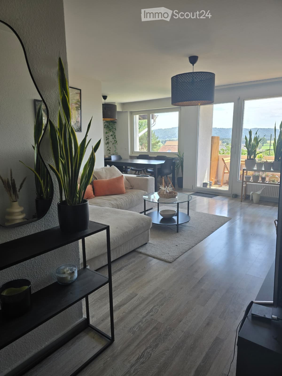 Modern living room with a light-colored sofa, coffee table, side table, and dining area visible through an open doorway. The room features large windows with a scenic view and is decorated with potted plants and a mirror on the wall.