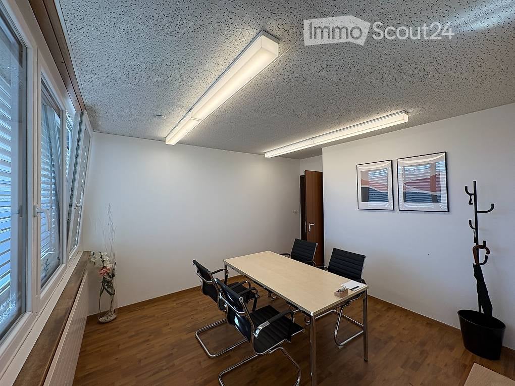 Bright, modern office space with wooden flooring, large windows, a table surrounded by chairs, and framed pictures on the wall.