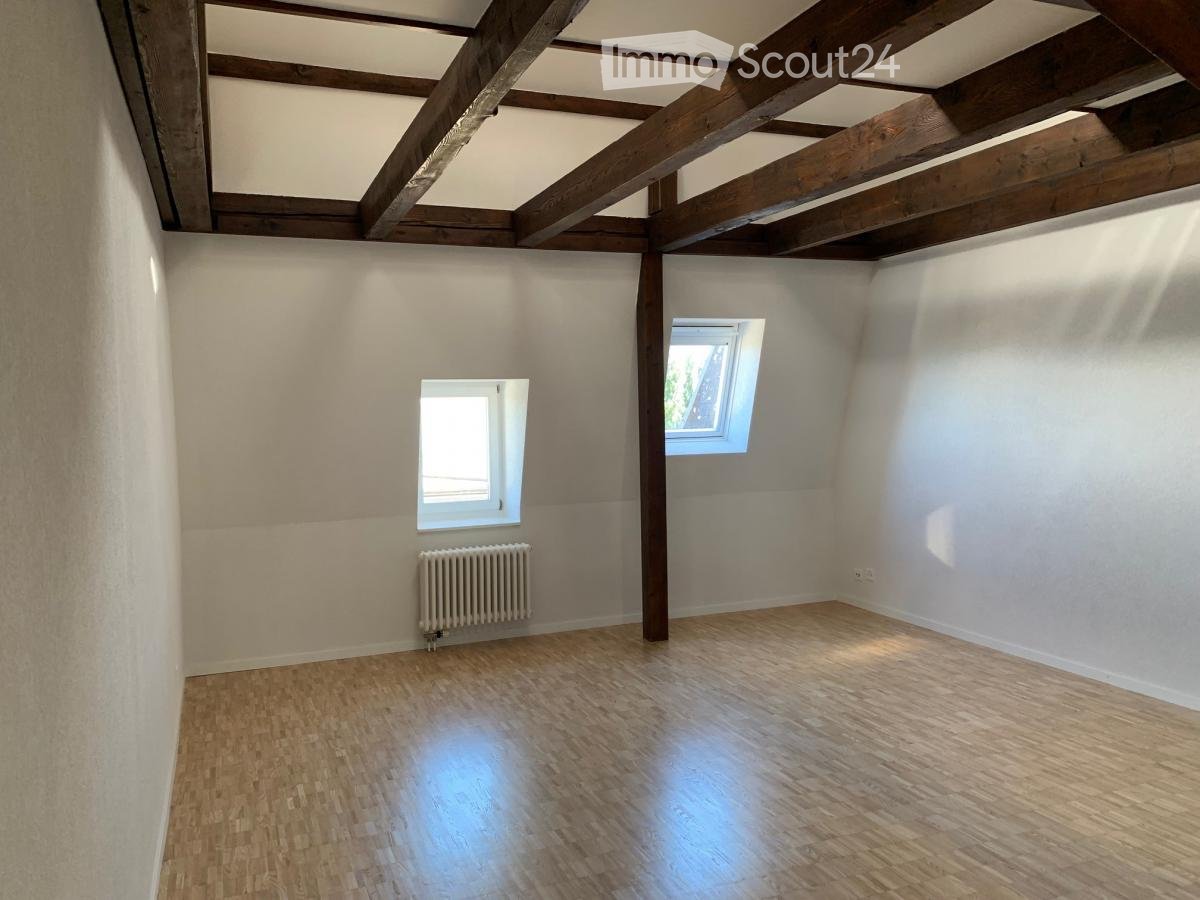Empty room with wooden floor, white walls, exposed wooden beams on the ceiling, two windows, and a radiator.