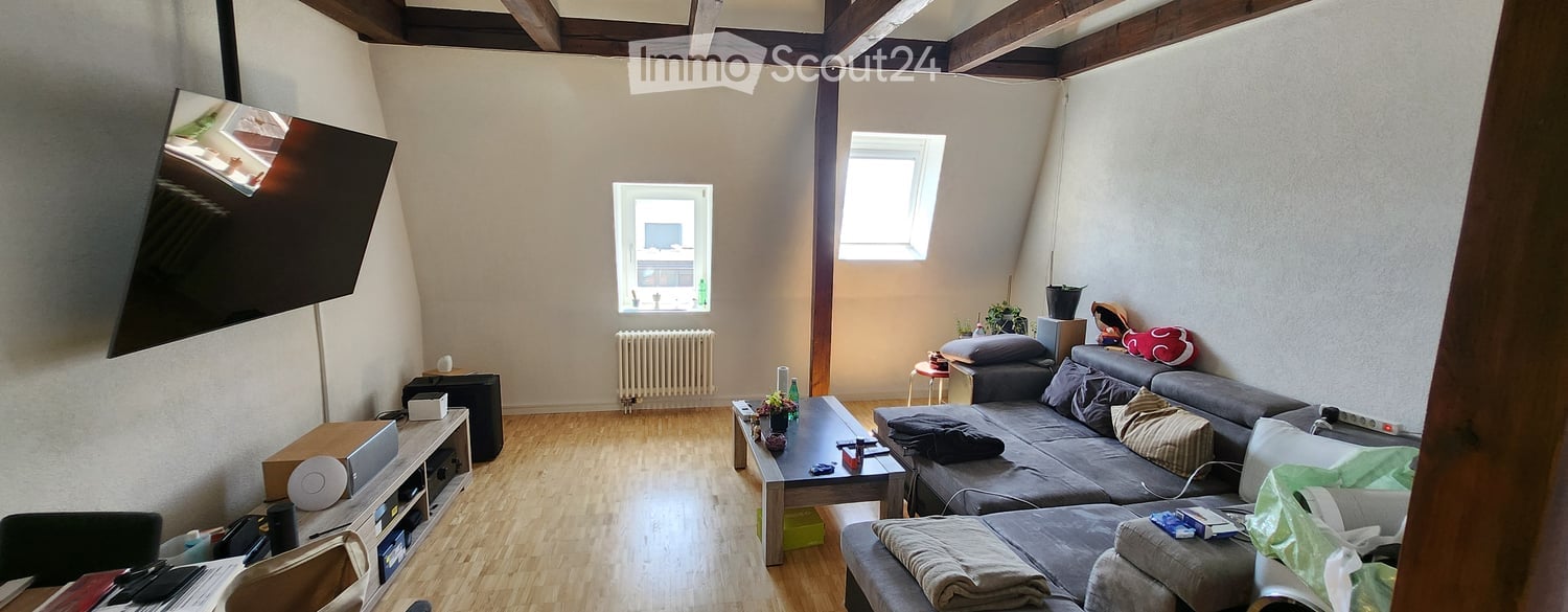 Living room with wooden flooring, exposed wooden beams on the ceiling, large windows, grey sofa, coffee table, TV stand, radiator, and small stool.