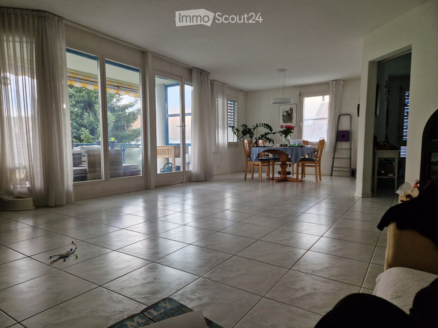 Bright dining area with large windows and sliding glass doors leading to a balcony. The room features a dining table with chairs, a hanging light fixture, and a fireplace on the right side.
