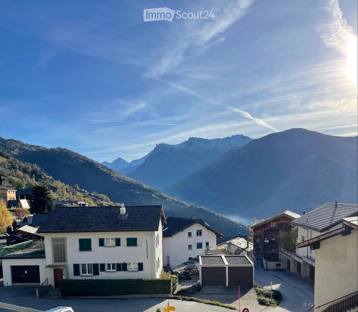 A two-story house with a dark roof and white walls, featuring multiple windows and a garage, situated in a picturesque mountain village setting.