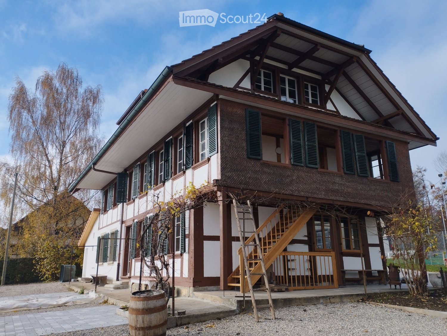 Two-story traditional house with white and brown exterior, green shutters, wooden staircase leading to a porch, and a wooden barrel in the foreground.