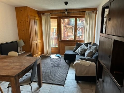 A cozy living room with wooden furniture, a sofa, coffee table, and a dining table. Large windows offer a view of the balcony and surrounding area.
