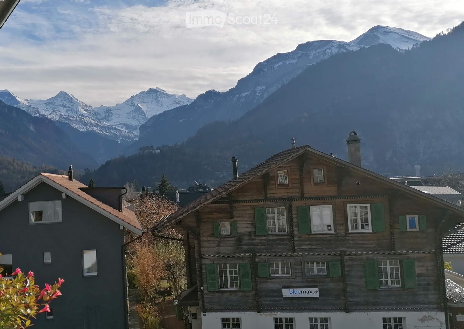 Traditional wooden house with green shutters and a red-tiled roof, situated in a mountainous area with snow-capped peaks in the background.