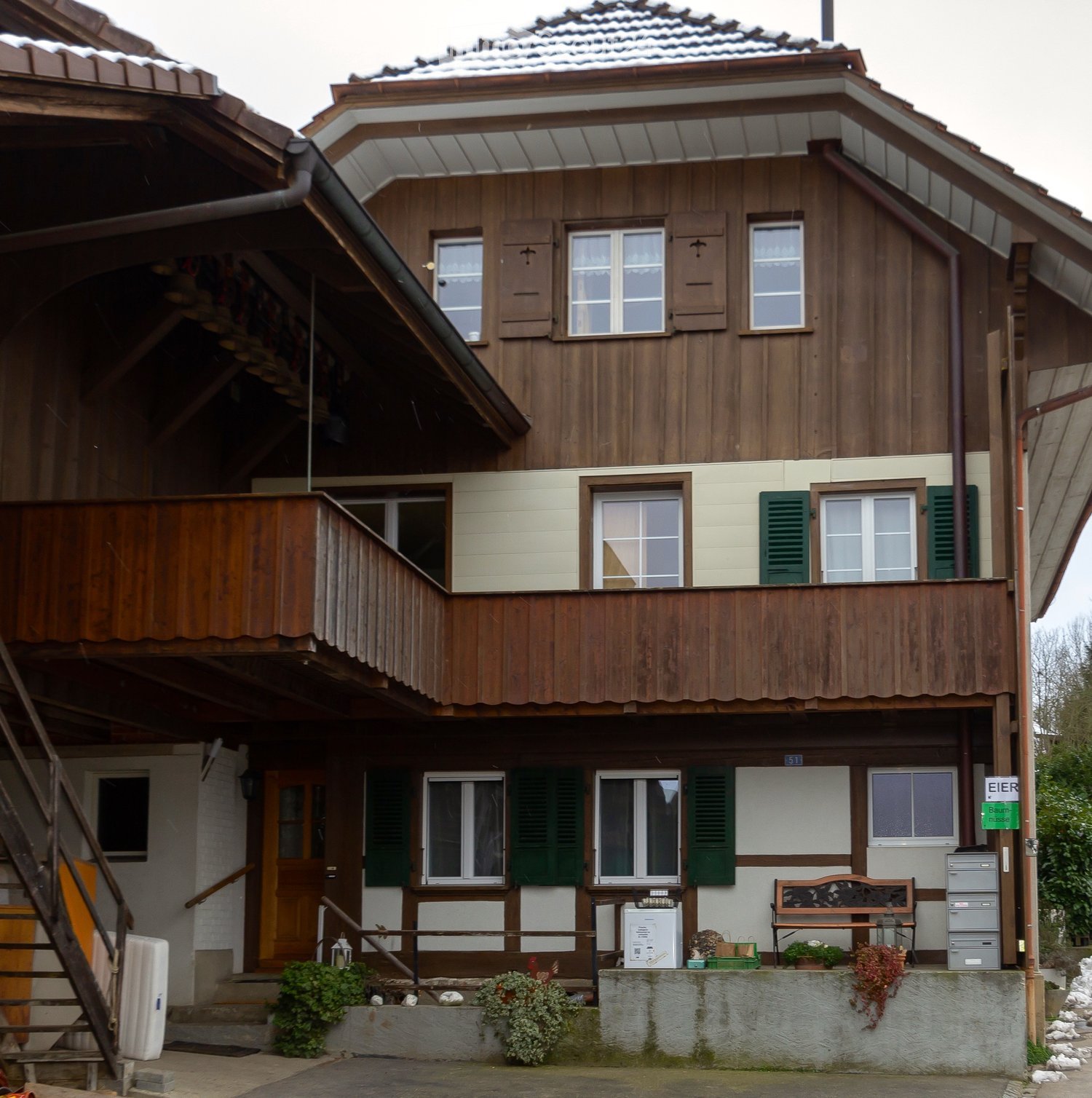 Two-story wooden house with green shutters, a wooden balcony, and a small porch area with a bench and potted plants.