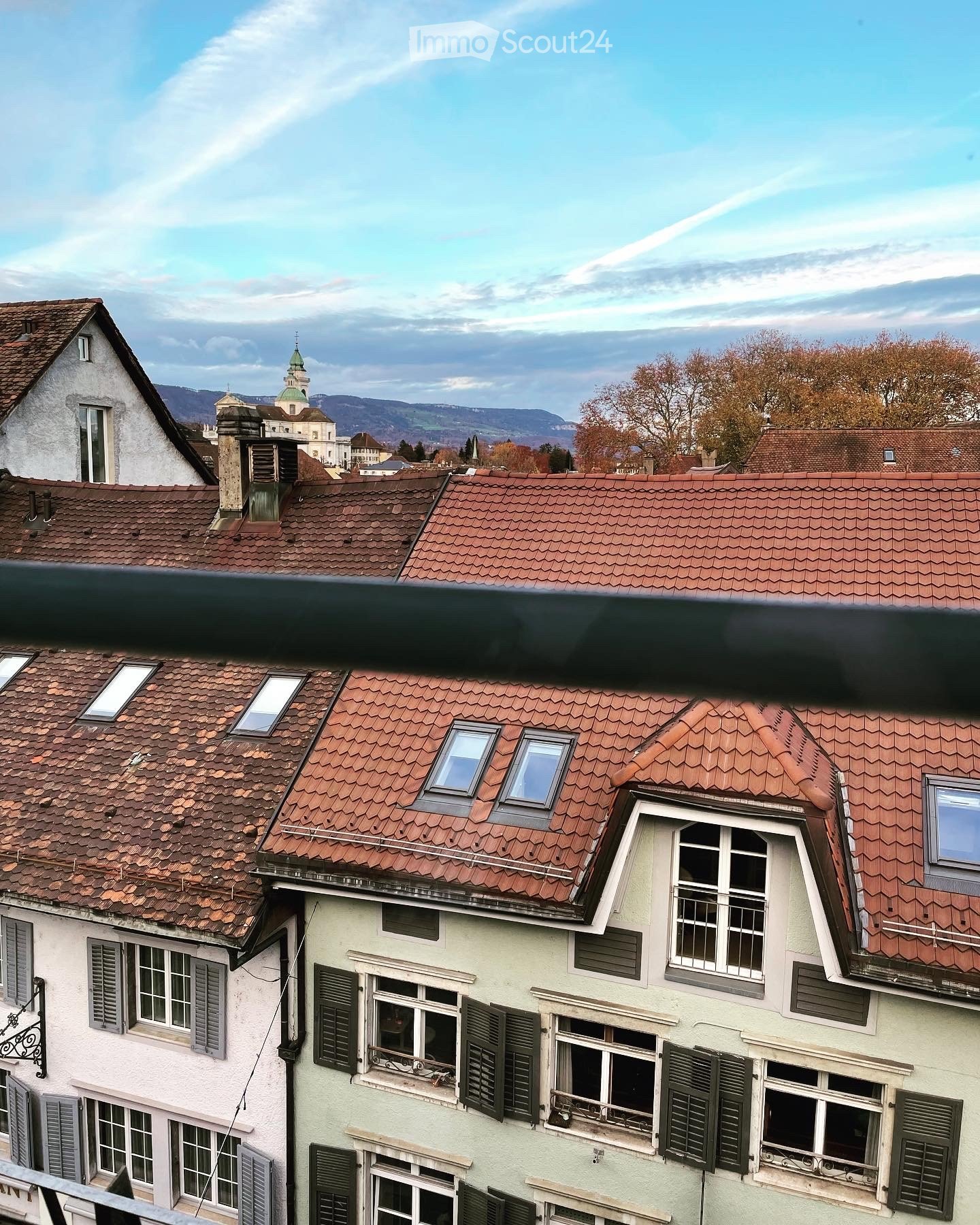 A multi-story residential building with red-tiled roofs, white walls, and multiple windows with shutters. The scene includes a distant church spire and mountains in the background.