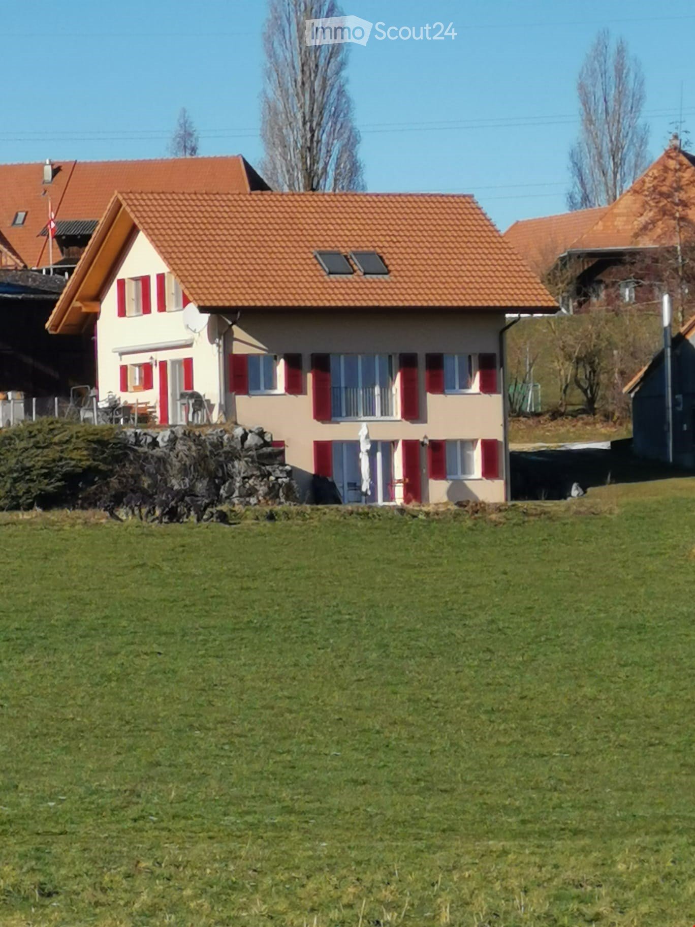 Two-story house with red-tiled roof, beige walls, red shutters, and a small balcony. The house is surrounded by a grassy lawn and a stone wall.