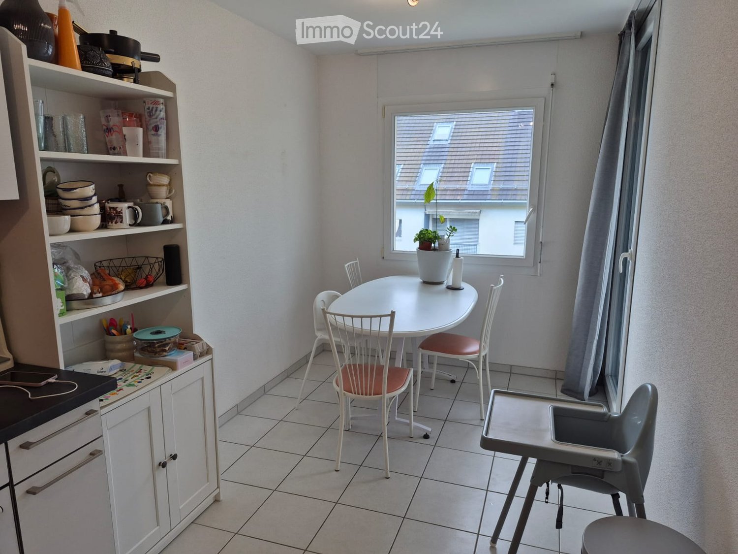 A small, well-lit room with a round white table and four chairs, a shelf with dishes and cups, a potted plant on the windowsill, and a baby chair in the corner.