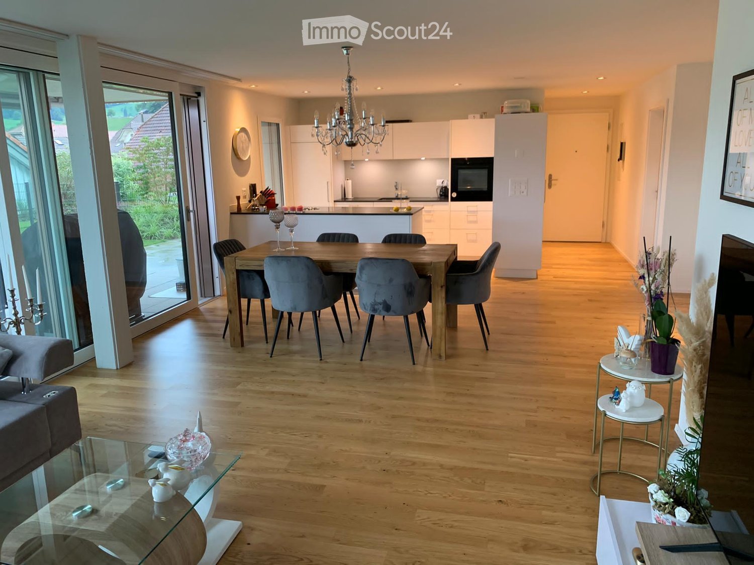 Open-plan living and dining area with light wooden flooring, a modern sofa, a glass coffee table, a dining table with six chairs, and a fully equipped kitchen with white cabinets and a microwave. The space features a chandelier, side tables with decorativ