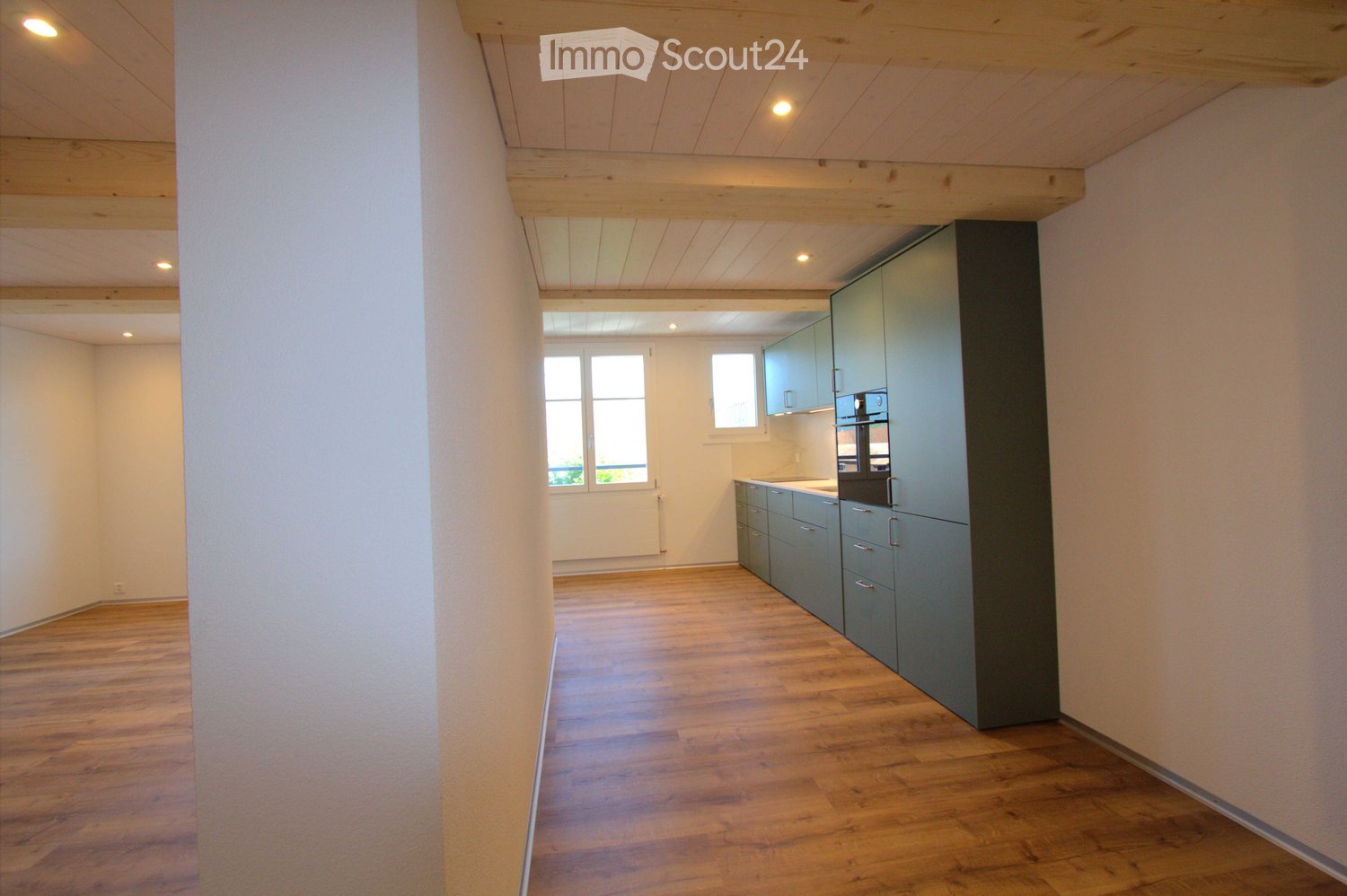 Open-plan space with wooden flooring, exposed wooden beams on the ceiling, and grey kitchen cabinets along one wall. The room is empty, with no furniture or appliances visible.