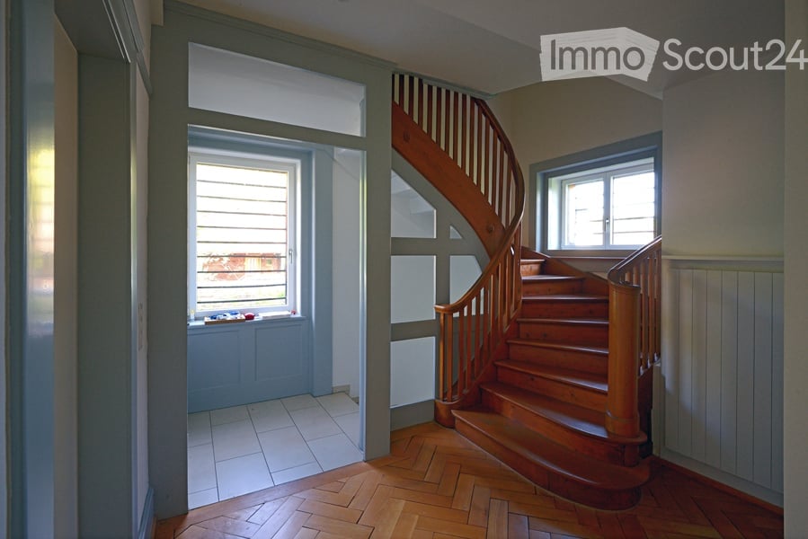 Spacious hallway with wooden flooring, a wooden staircase leading upwards, and large windows providing natural light. The walls are painted in neutral tones, and there is a small window seat with a few decorative items on the left side.