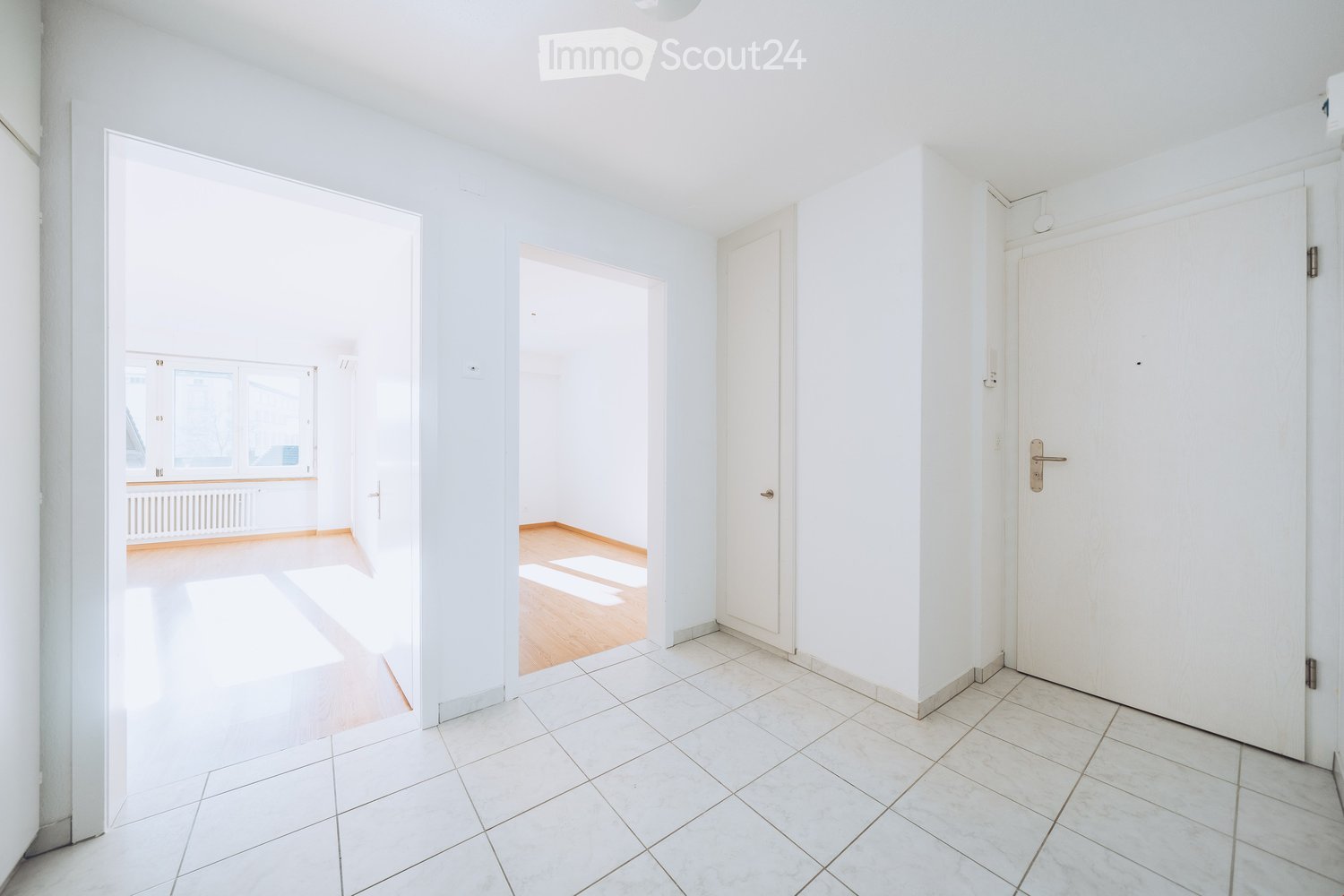 Empty room with white walls, tiled floor, and multiple doors leading to other rooms. Natural light streams in from a window in the adjacent room.