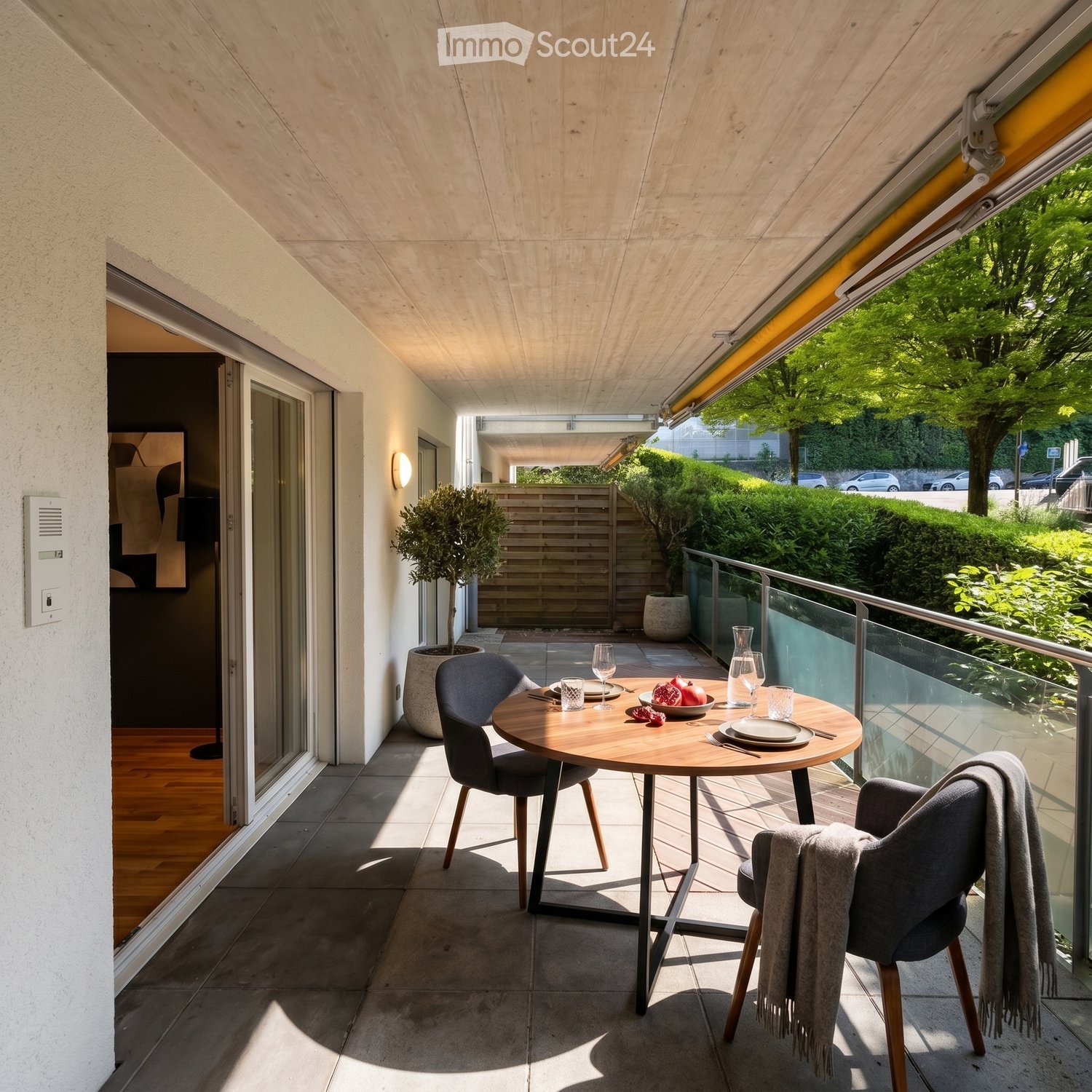 Modern balcony with a round wooden table, two grey chairs, wine glasses, plates, and a bowl of pomegranate. The balcony has a glass railing and is partially covered by a retractable awning. Potted plants are placed nearby.