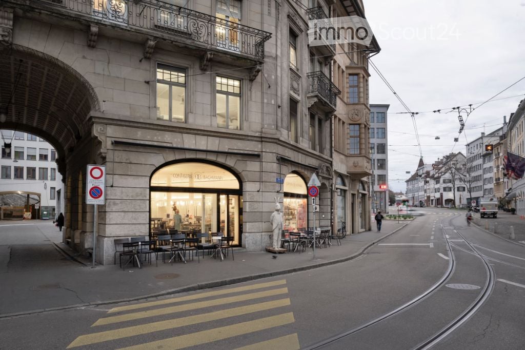 Urban street view with a multi-story building featuring large windows, balconies, and a storefront with outdoor seating. The street includes traffic signs, a pedestrian crossing, and a tram line.