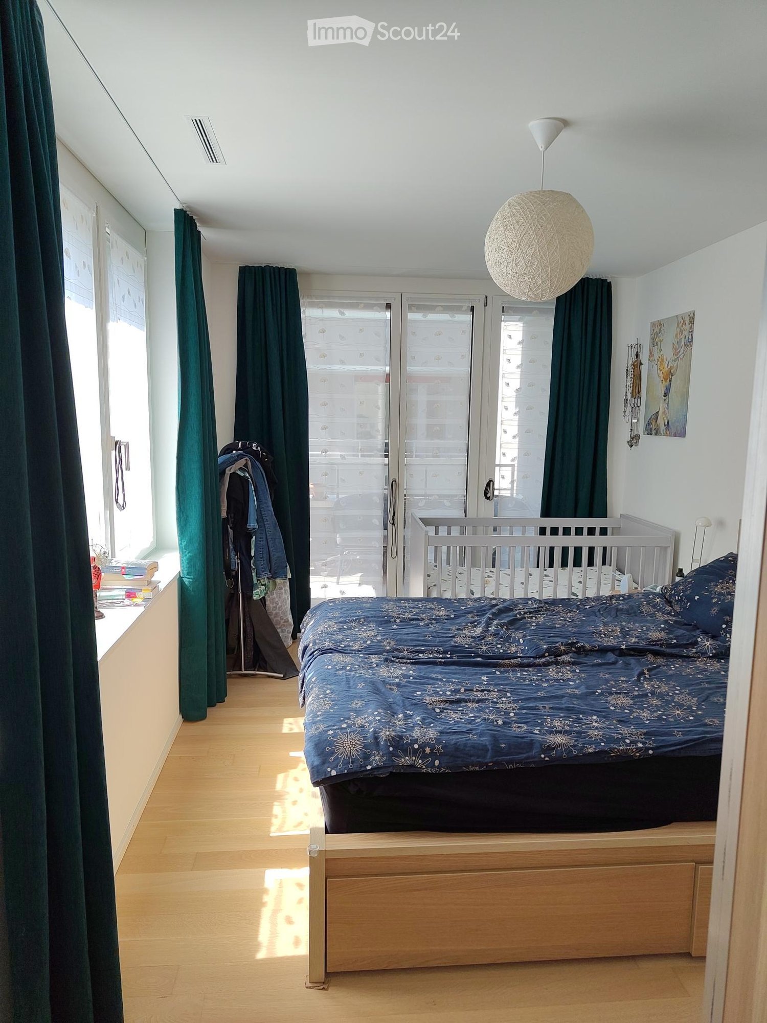 Bright bedroom with wooden flooring, large window with green curtains, bed with blue bedding, hanging lamp, and a painting on the wall.