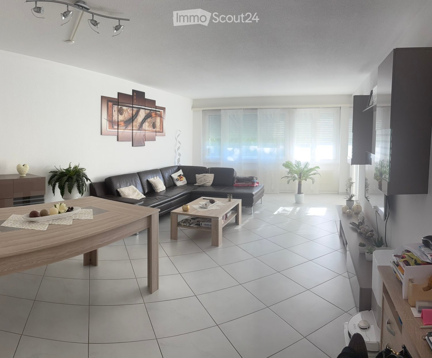 Modern living room with white tiled flooring, large windows, a black leather sectional sofa, a wooden coffee table, a dining table, sideboard, and decorative plants.