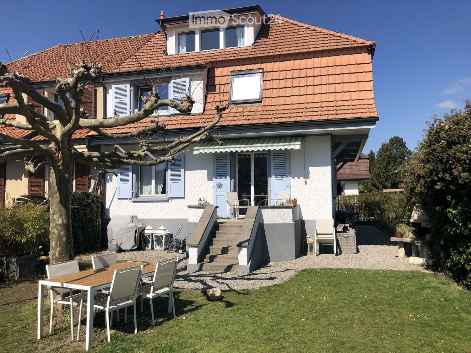 A two-story house with a red-tiled roof and white walls, featuring a well-maintained garden with a wooden table and chairs, steps leading to a covered patio area, and various outdoor furniture.
