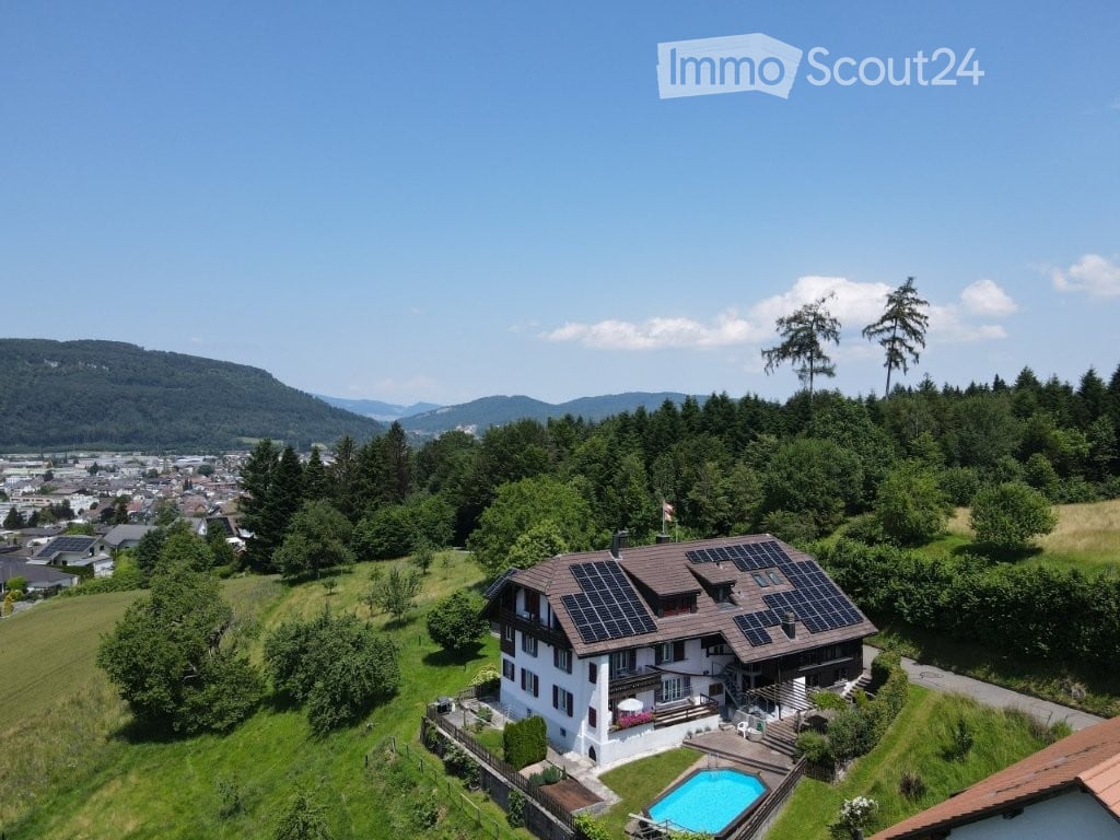 A two-story house with solar panels on the roof, surrounded by a well-maintained garden and a swimming pool. The house is situated on a hillside with a scenic view of the valley and mountains in the background.