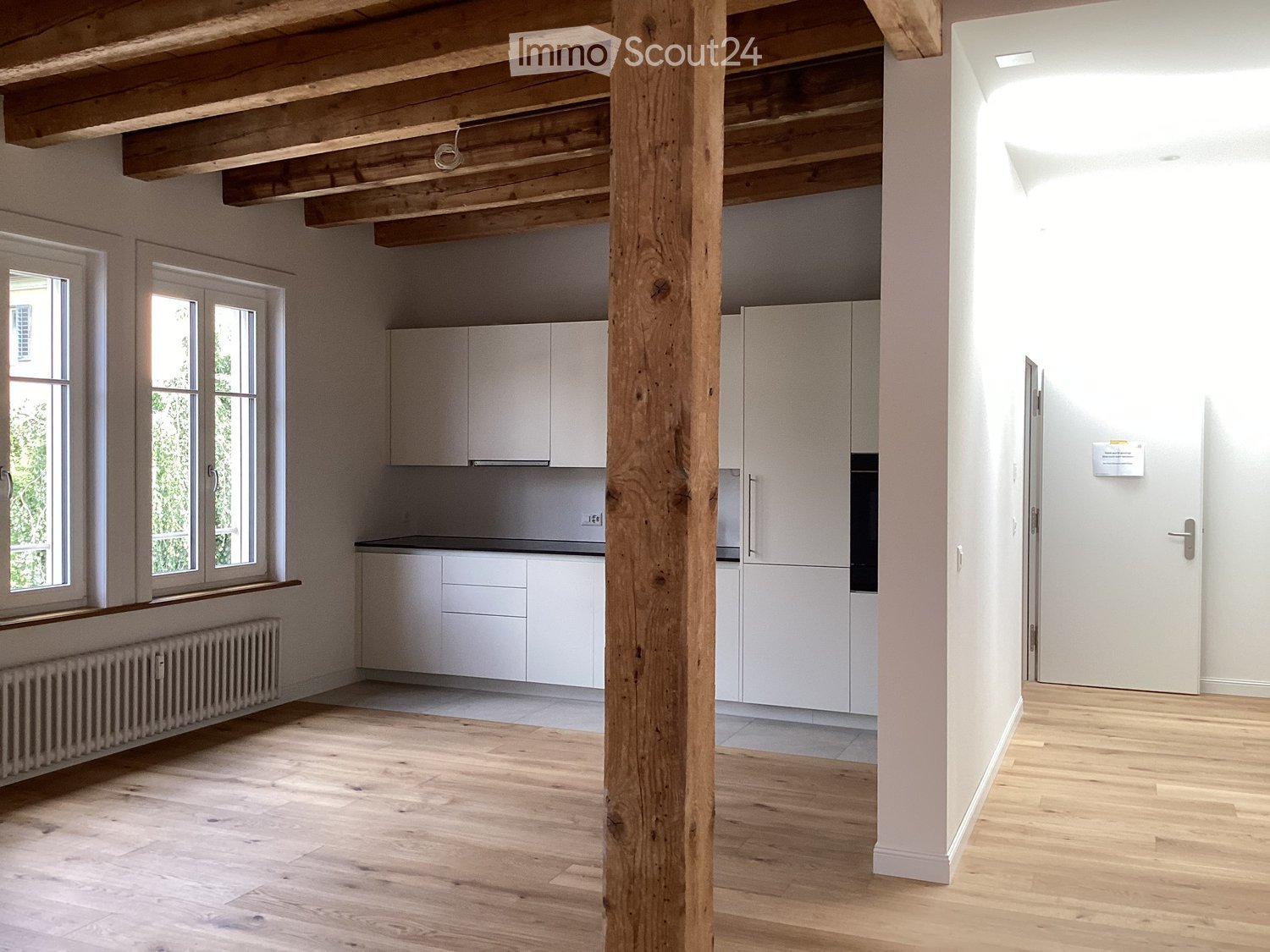 Empty room with wooden floor, white walls, exposed wooden beams, white kitchen cabinets, and windows with white frames.