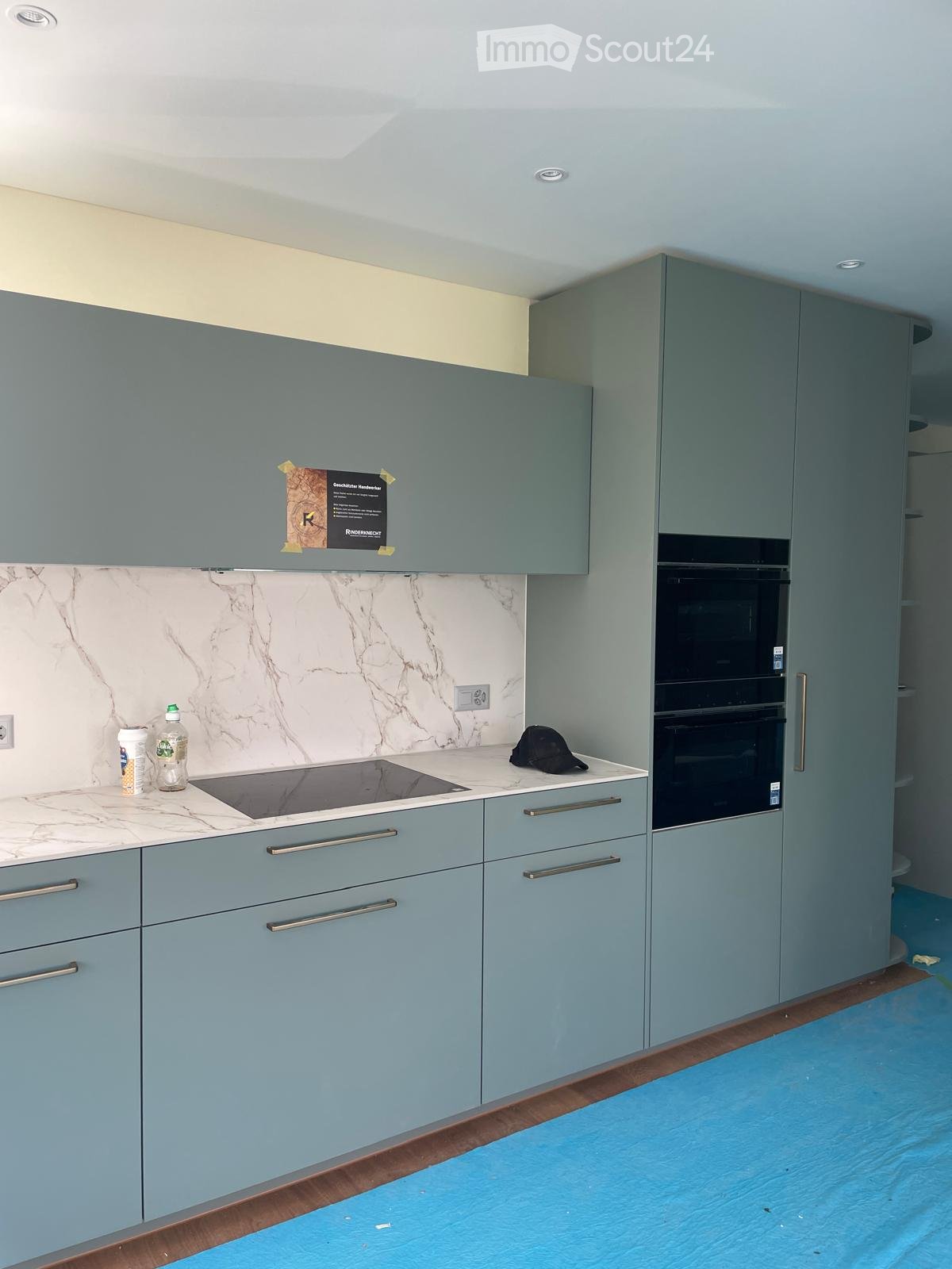 Modern kitchen with light blue cabinets, marble backsplash, induction stove, and double oven. Countertop has a few items including a bottle and a hat.
