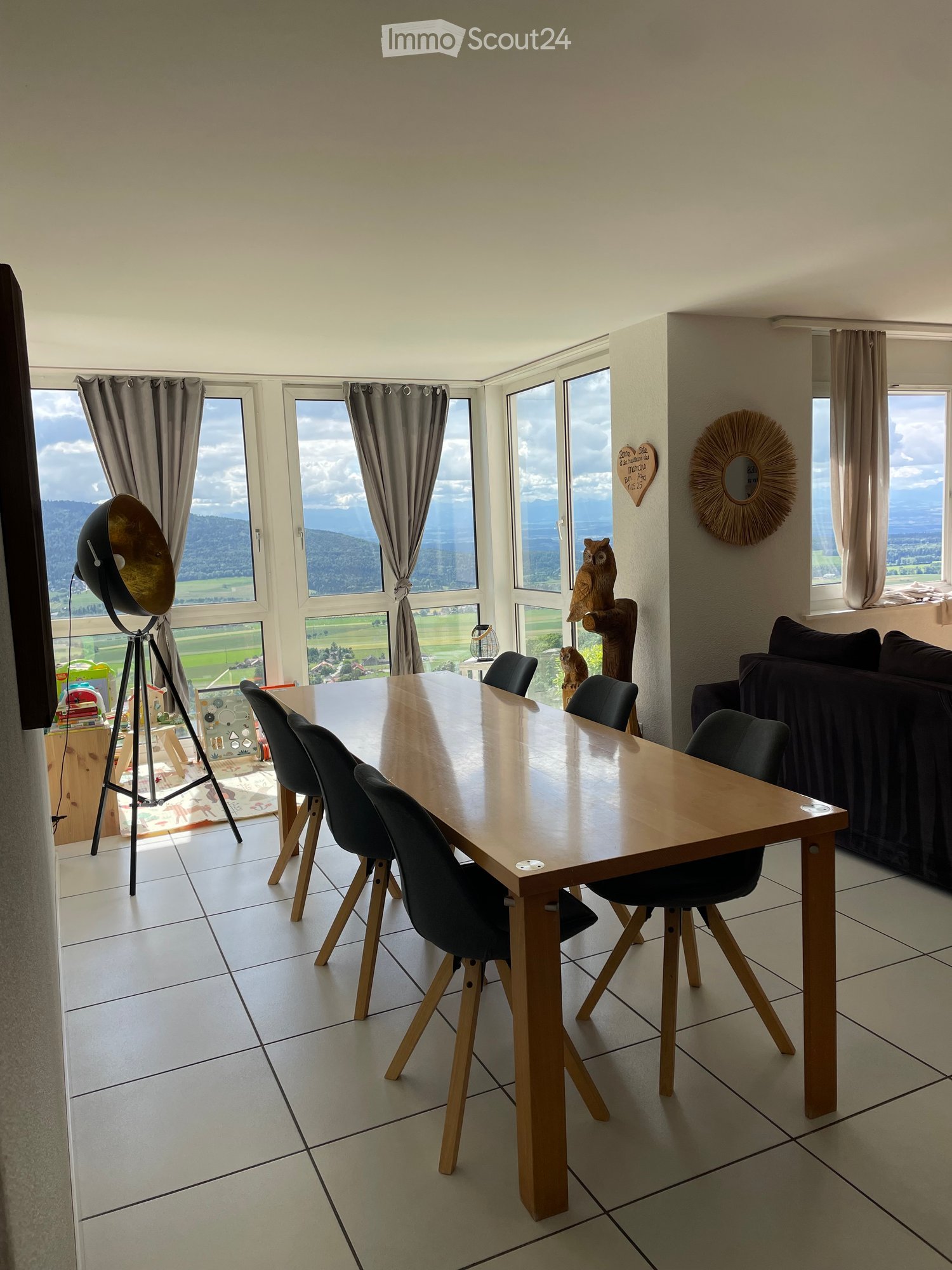 Bright dining area with large windows offering a scenic view, featuring a wooden dining table, black chairs, a sofa, and decorative items including a mirror and lamp.