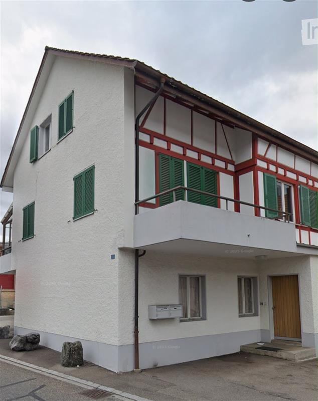 Two-story building with white walls, green shutters, and a balcony on the upper floor.