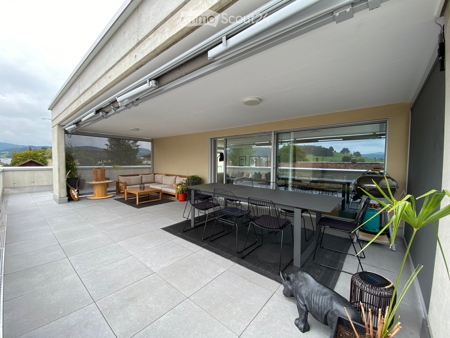 Modern balcony with tiled floor, large glass windows, sofa, coffee table, dining area, barbecue, and scenic mountain view.