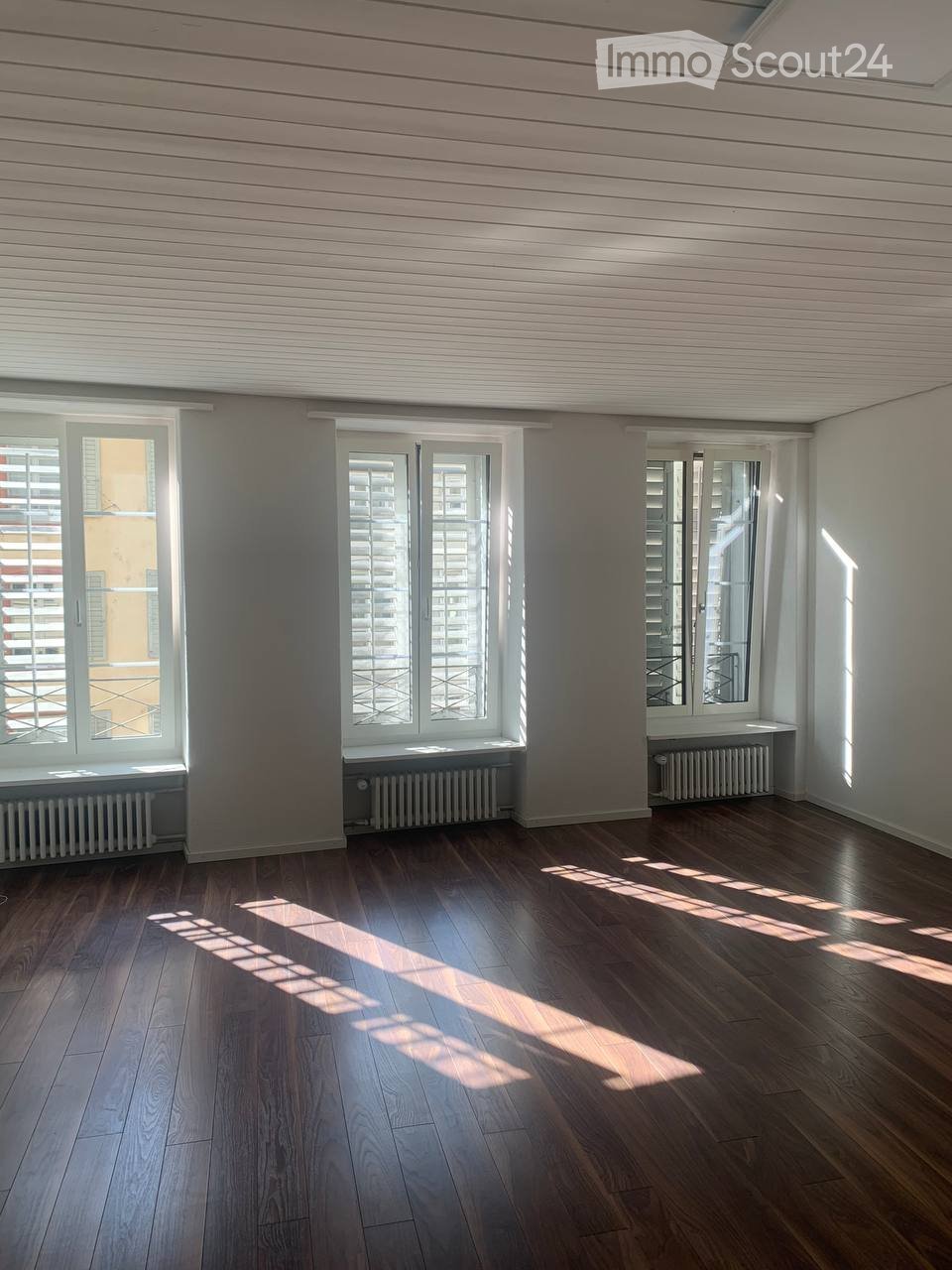Empty room with wooden flooring, large windows with shutters, radiators, and natural light streaming in.