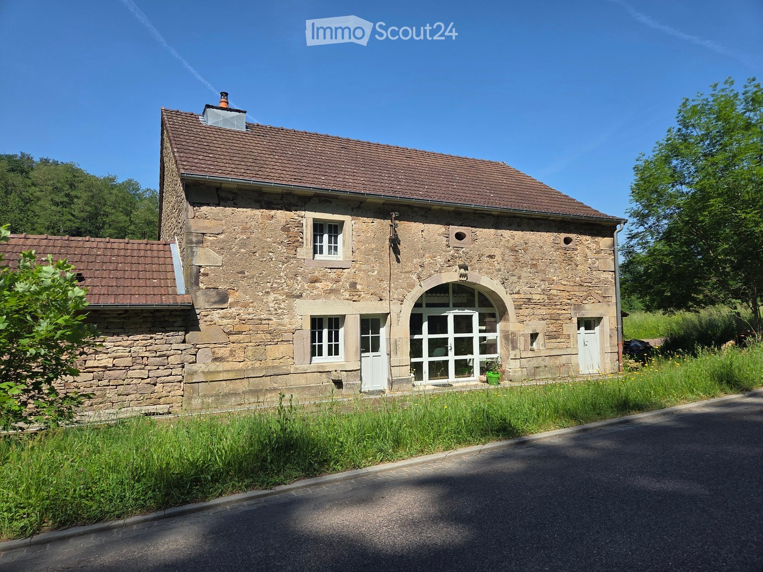 Two-story stone house with a tiled roof, multiple windows, and a garage entrance. Surrounded by greenery and a paved road.