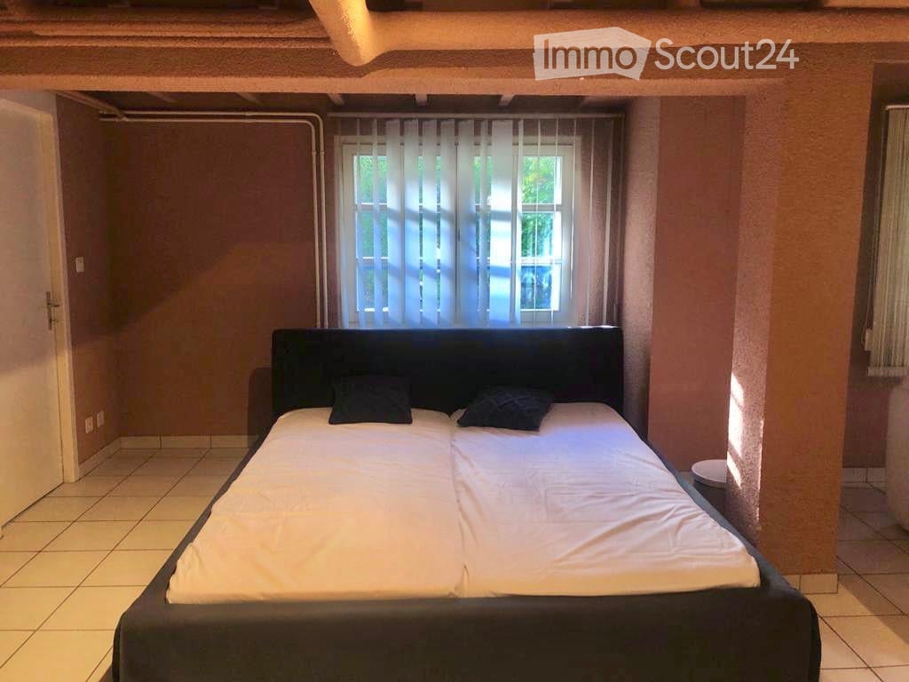 A bedroom with a double bed, two pillows, and a window with sheer curtains. The room has tiled flooring and warm-colored walls.