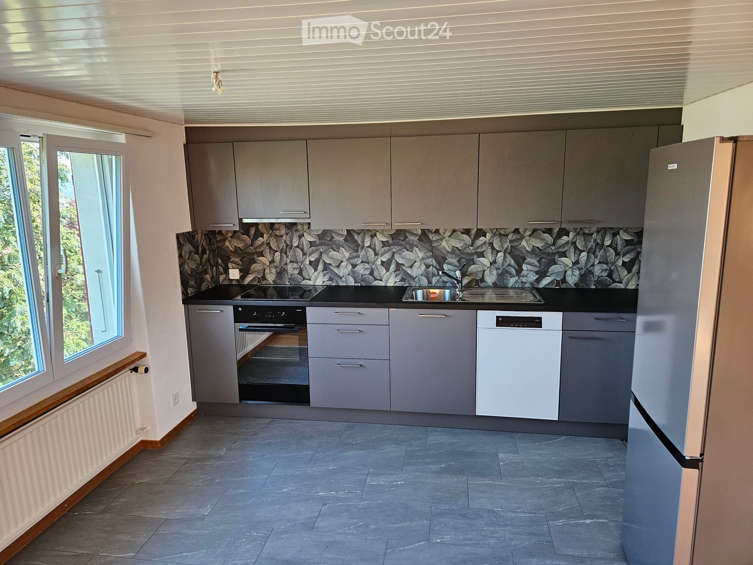 Modern kitchen with grey cabinets, black countertops, stainless steel appliances, and a decorative backsplash featuring leaf patterns. Large windows provide natural light.