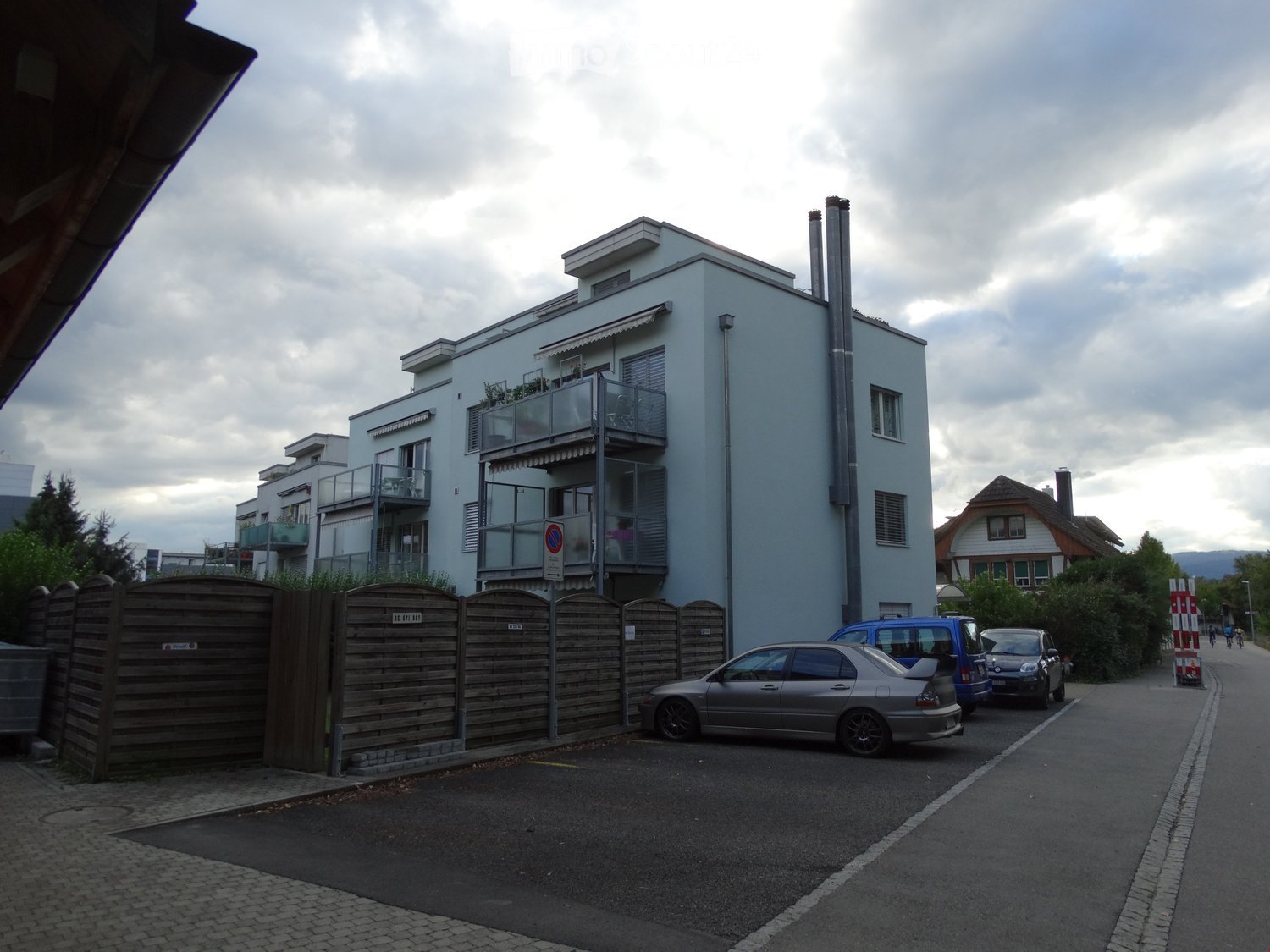 Modern multi-story building with balconies, surrounded by a wooden fence and parked cars.