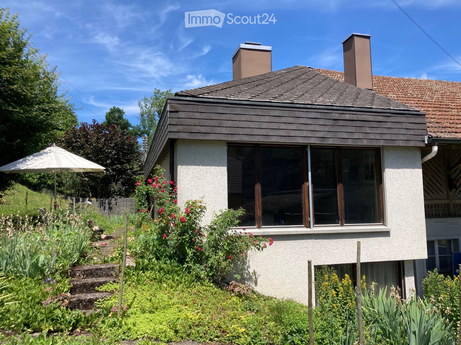 Single-story house with a modern design, featuring a raised ground floor, large windows, and a well-maintained garden with colorful flowers and an umbrella.