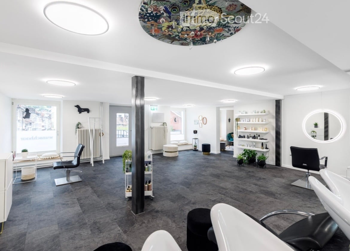 Modern, well-lit salon with sleek black chairs, white sinks, round mirrors, shelves with products, and decorative plants.