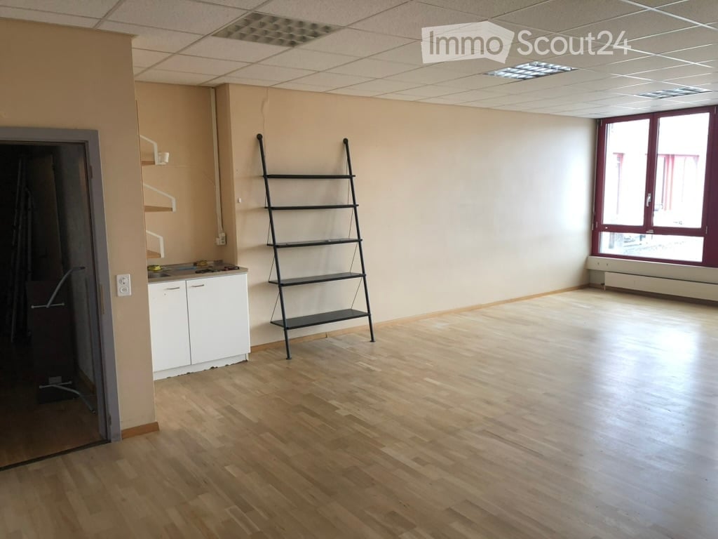 Empty room with wooden flooring, white walls, and a small kitchenette area with cabinets and a countertop. A tall, black metal shelving unit is positioned against one wall, and a window with red frames is visible on the right side.
