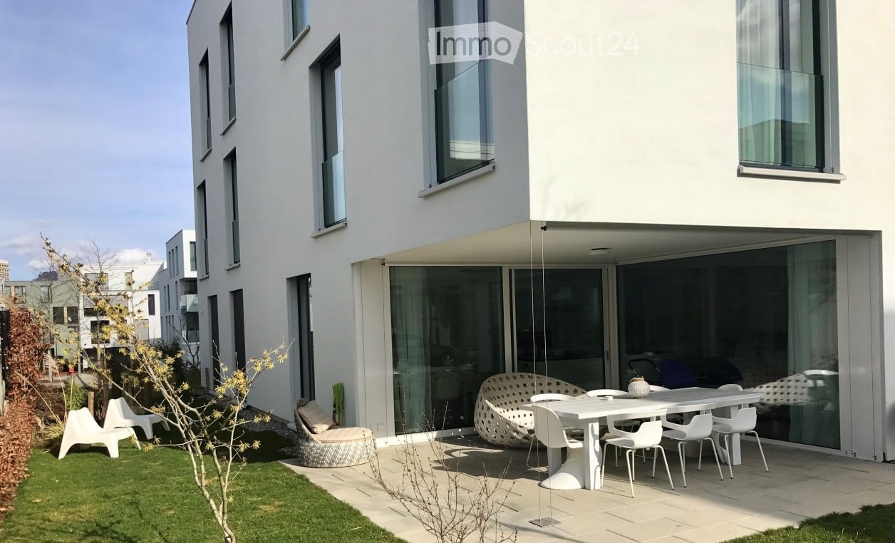 Modern outdoor patio area attached to a contemporary white building, featuring a white dining table with chairs, a white sofa, and a small grassy area with a tree.