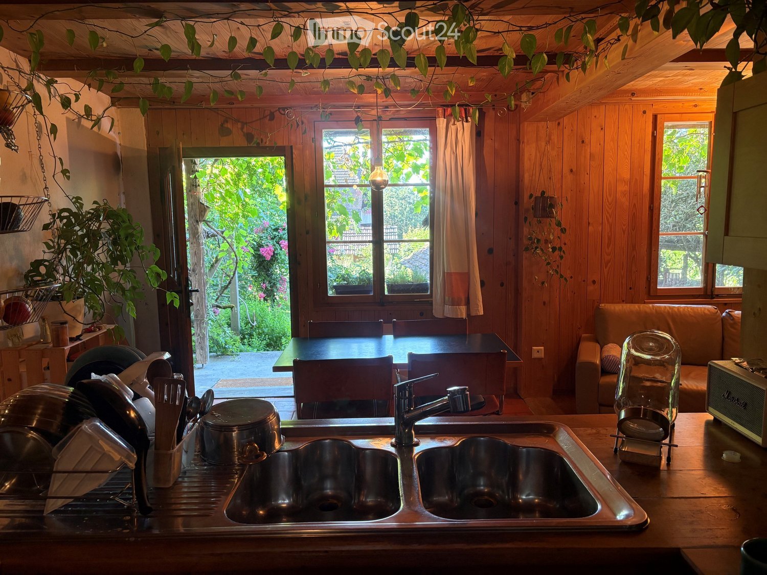 Cozy kitchen with wooden walls and ceiling, featuring a double sink, dish rack, table, chairs, and a sofa near the window overlooking a garden.