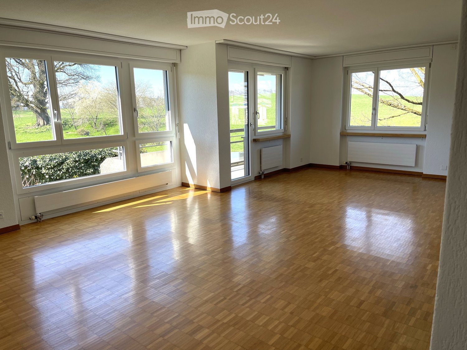 Empty room with wooden flooring, large windows, and radiators along the walls.