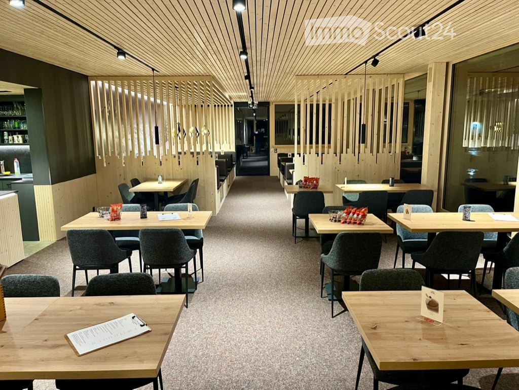 Modern restaurant interior with wooden tables, grey chairs, and pendant lighting. Menu and cups are placed on tables.