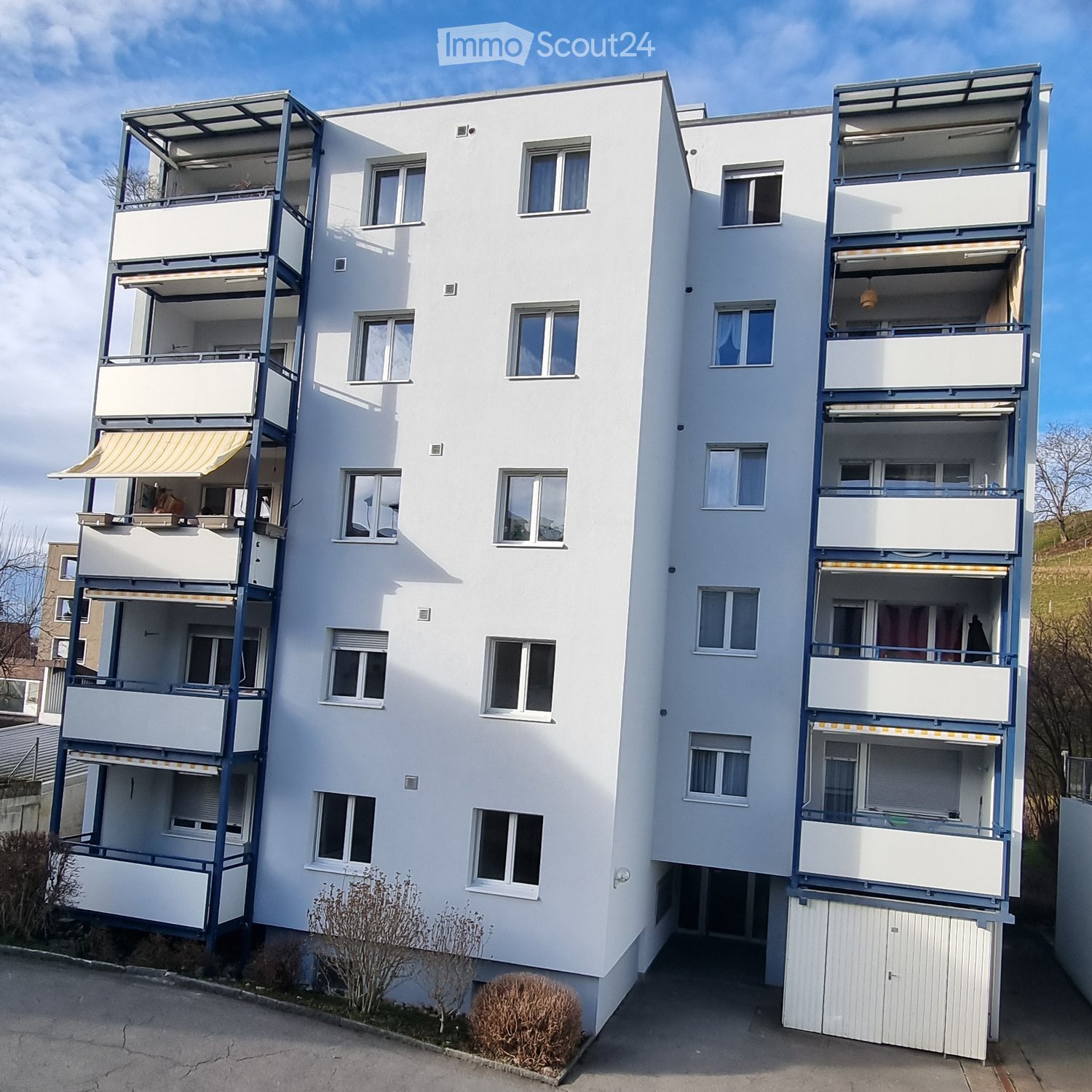 Modern multi-story apartment building with white facade, multiple balconies, and large windows. Features include a covered entrance and a small garden area at the base.
