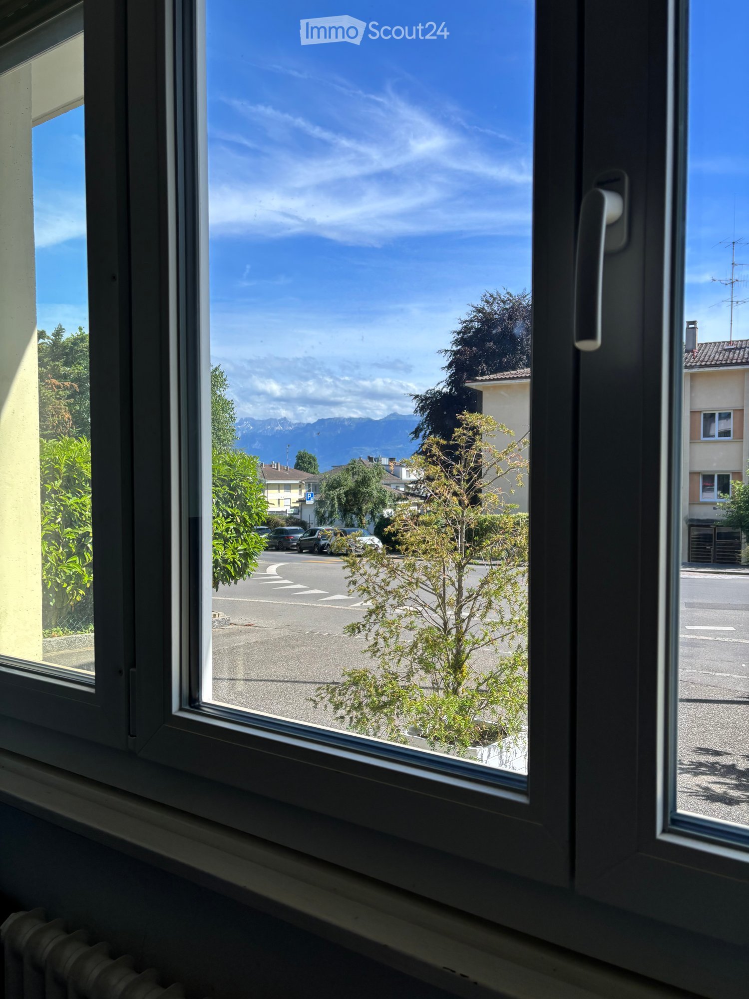 View from a window showing a street, parked cars, trees, and distant mountains under a clear blue sky.