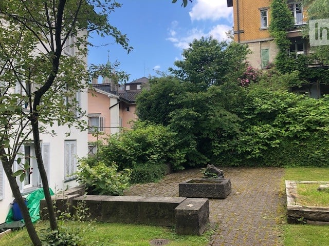 A small, enclosed garden area with gravel pathways, stone benches, and lush greenery, surrounded by residential buildings.