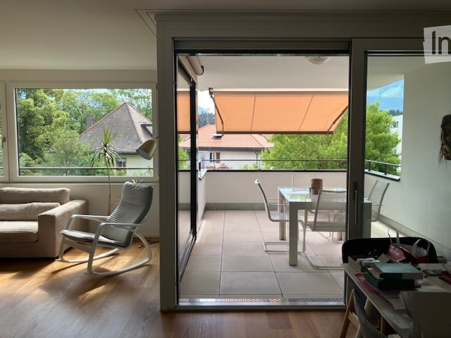 Bright living room with large windows and sliding glass doors leading to a balcony. Features a sofa, armchair, dining table with chairs, and a small shelf with books.